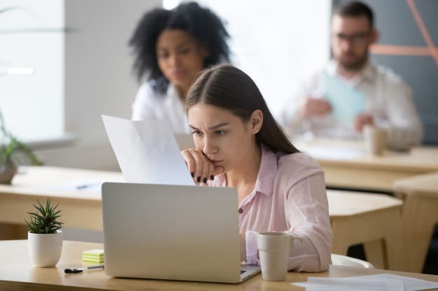 Bain online test. Image shows a woman sitting at a table with her laptop open and studying a sheet of paper.