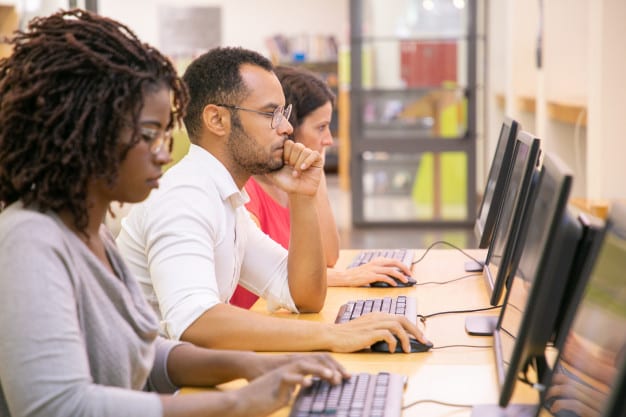 Bain online multiple choice test. Image shows a row of 3 people taking tests on computers at a college library.